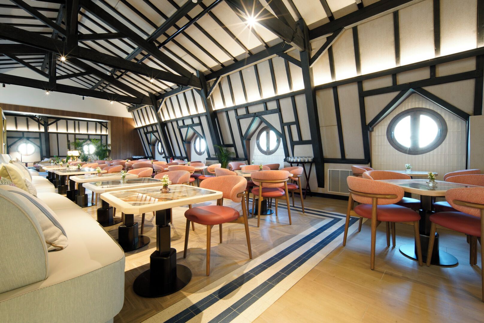 Un restaurante con techos altos, vigas a la vista y ventanas redondas. Mesas y sillas dispuestas para comer.