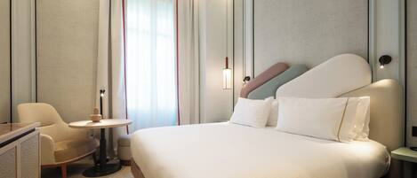 A hotel room with a bed, a desk, a chair, a small table, and a window with curtains.