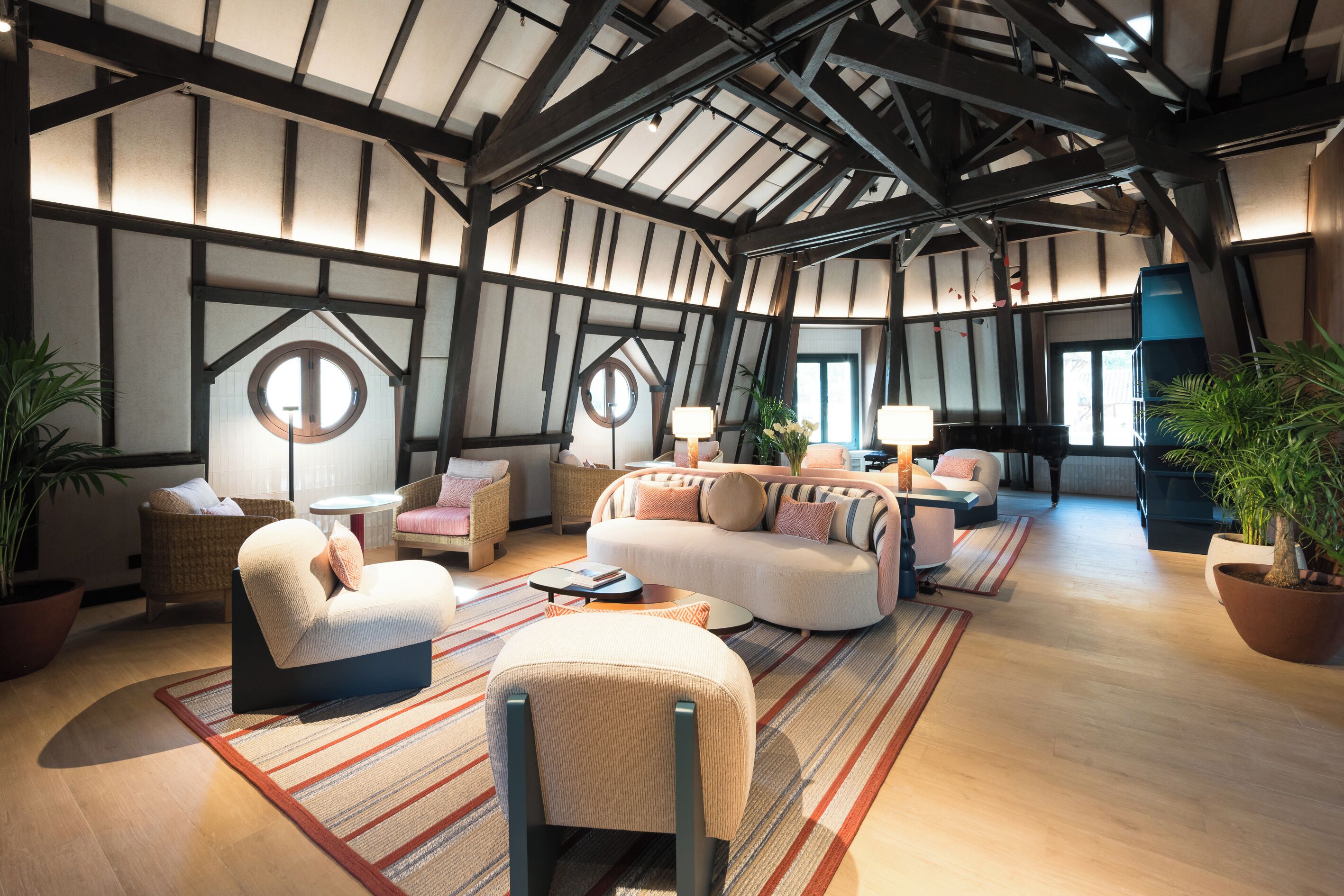 A spacious living area with a high ceiling, exposed beams, and large windows. There are multiple seating areas with sofas and armchairs, and a piano in the background.