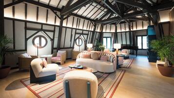 A spacious living area with a high ceiling, exposed beams, and large windows. There are multiple seating areas with sofas and armchairs, and a piano in the background.