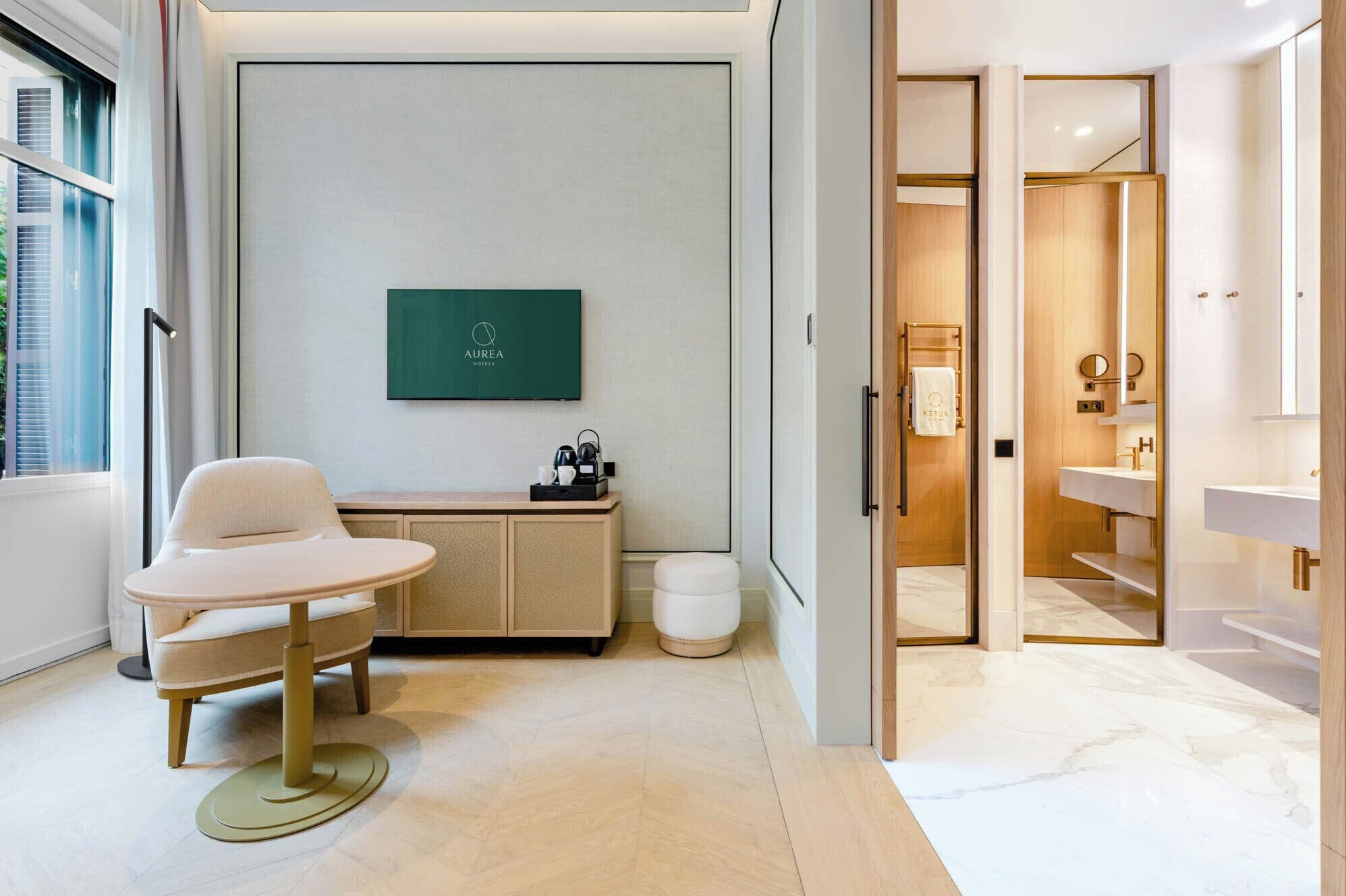 Una habitación de hotel moderna con un diseño minimalista, que incluye una zona de estar con una mesa y sillas, un escritorio pequeño con cafetera, un espejo grande y un baño independiente con tocador y lavabo.