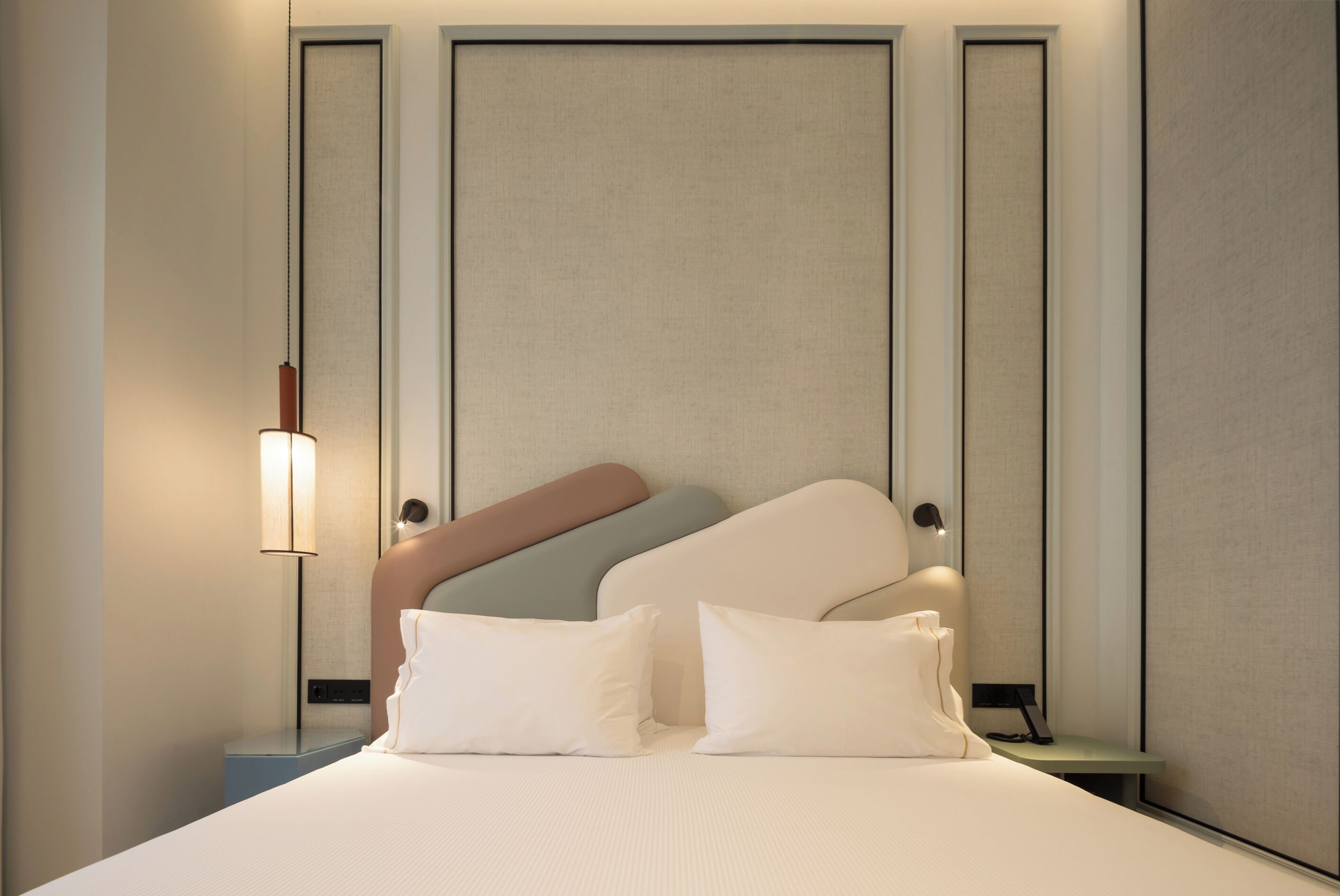 Una habitación de hotel ordenada con una cama grande, dos mesitas de noche, una lámpara de pared y un cabecero con varios colores de almohadas.