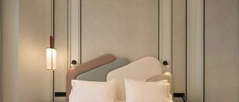 Una habitación de hotel ordenada con una cama grande, dos mesitas de noche, una lámpara de pared y un cabecero con varios colores de almohadas.