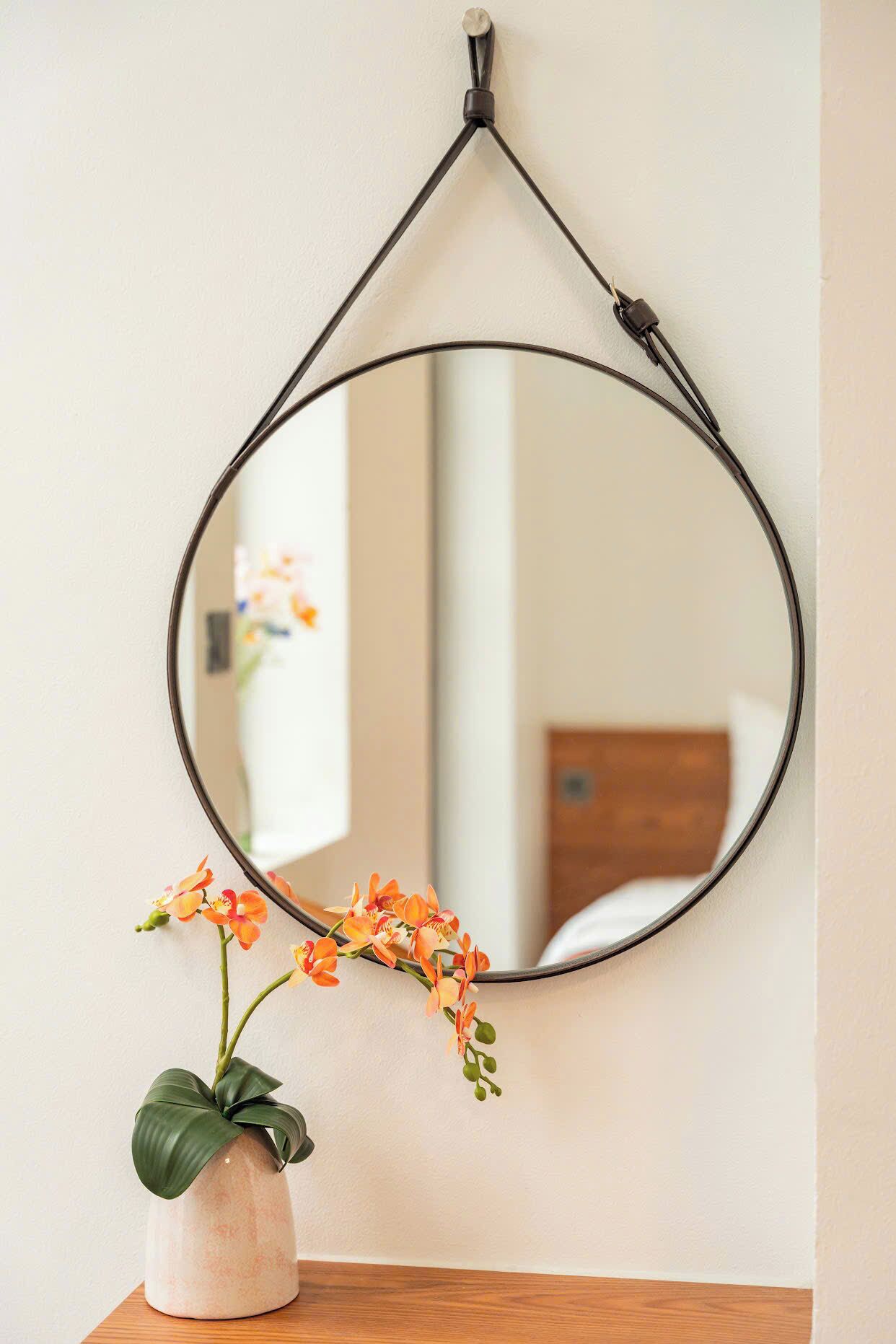 A round mirror with a black frame hanging on a wall, a small potted plant with orange flowers in front of it, and a reflection of a room with a bed and a window.