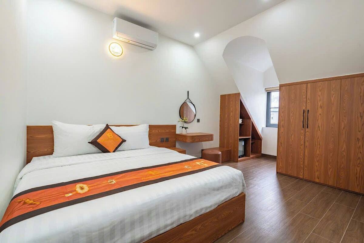 A modern bedroom with a large bed, a wooden wardrobe, a small desk, and a wall-mounted air conditioner.