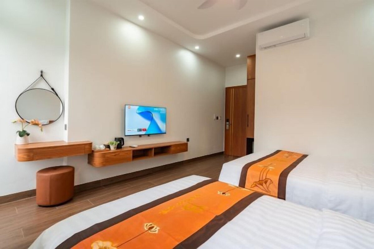 A modern hotel room with a flat-screen TV mounted on the wall, a wooden bench with storage, a trash bin, and two beds with orange bedspreads.