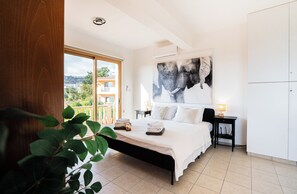 Quarto com cama, mesa de cabeceira, luminária, janela grande e vista para prédios.