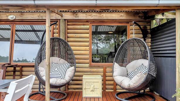 A wooden cabin with two hanging chairs with cushions, a wooden table, and a small cabinet. - Treetop Sanctuary Serene Cottage near Mount Lofty (Crafers West)