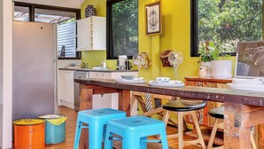 A compact kitchen with a dining area, featuring a wooden table, blue stools, and a view of greenery outside. - Treetop Sanctuary Serene Cottage near Mount Lofty (Crafers West)