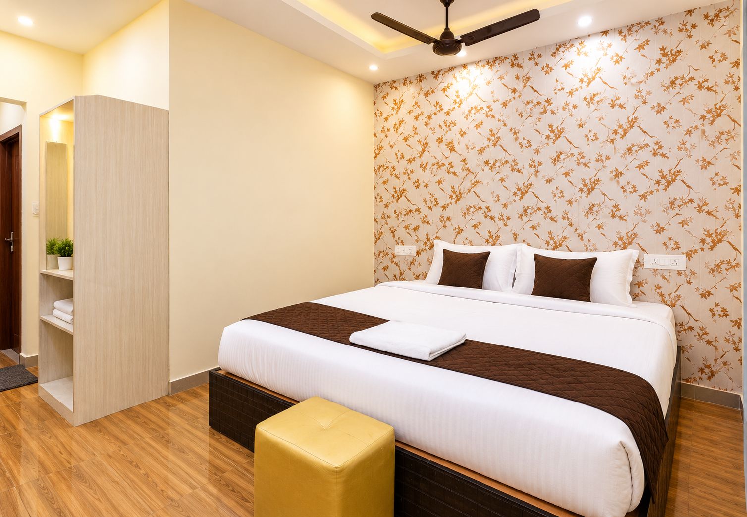 A hotel room with a bed, a ceiling fan, a wooden floor, a wall with floral wallpaper, and a small stool.