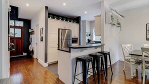 A modern kitchen with white cabinetry, stainless steel appliances, and a central island with bar stools. - Hôtel à la maison - Entre Mont et rivière (Orford)