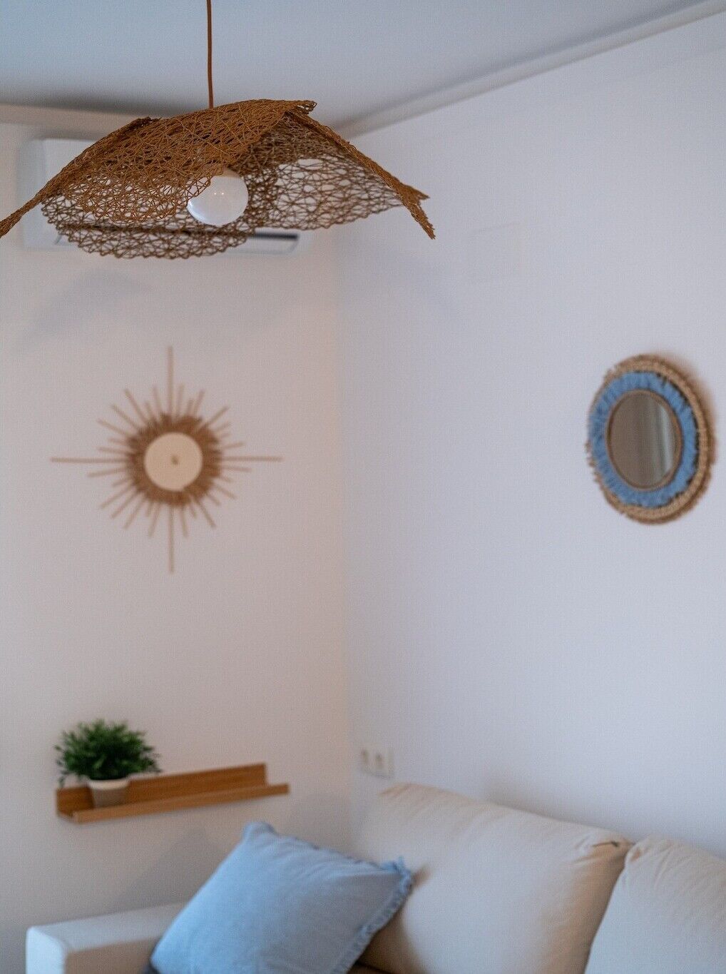 Ein Wohnzimmer mit einer hängenden gewebten Leuchte, einer Wanddekoration mit Sonnenelementen, einem Spiegel und einer kleinen Pflanze auf einem Regal.