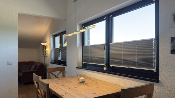Ein Essbereich mit einem Holztisch, Stühlen und einem Glas mit Kerze. Es gibt Fenster mit Jalousien und eine Deckenleuchte.