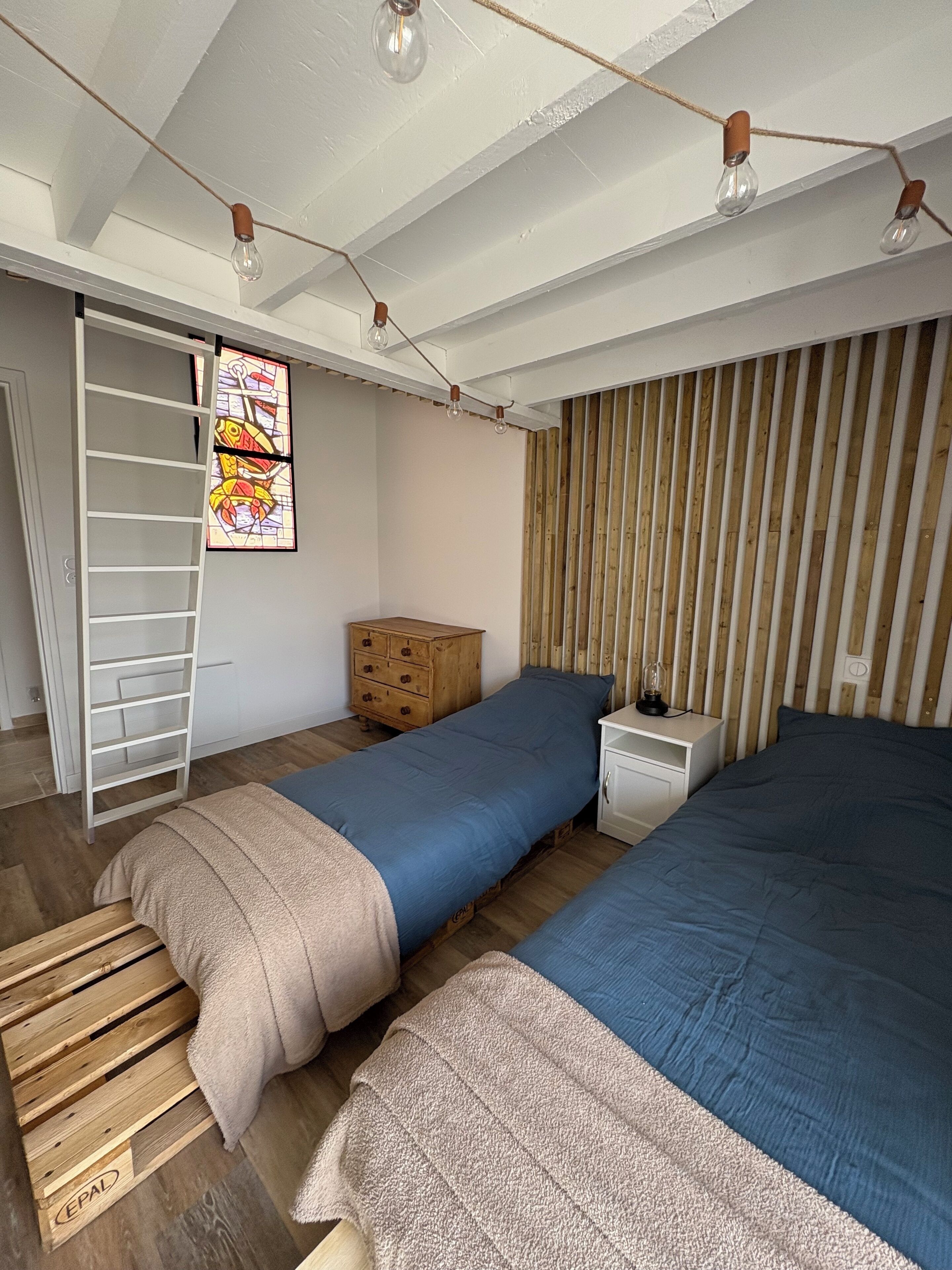 Une chambre avec deux lits, une commode en bois, une échelle et une fenêtre à vitraux.