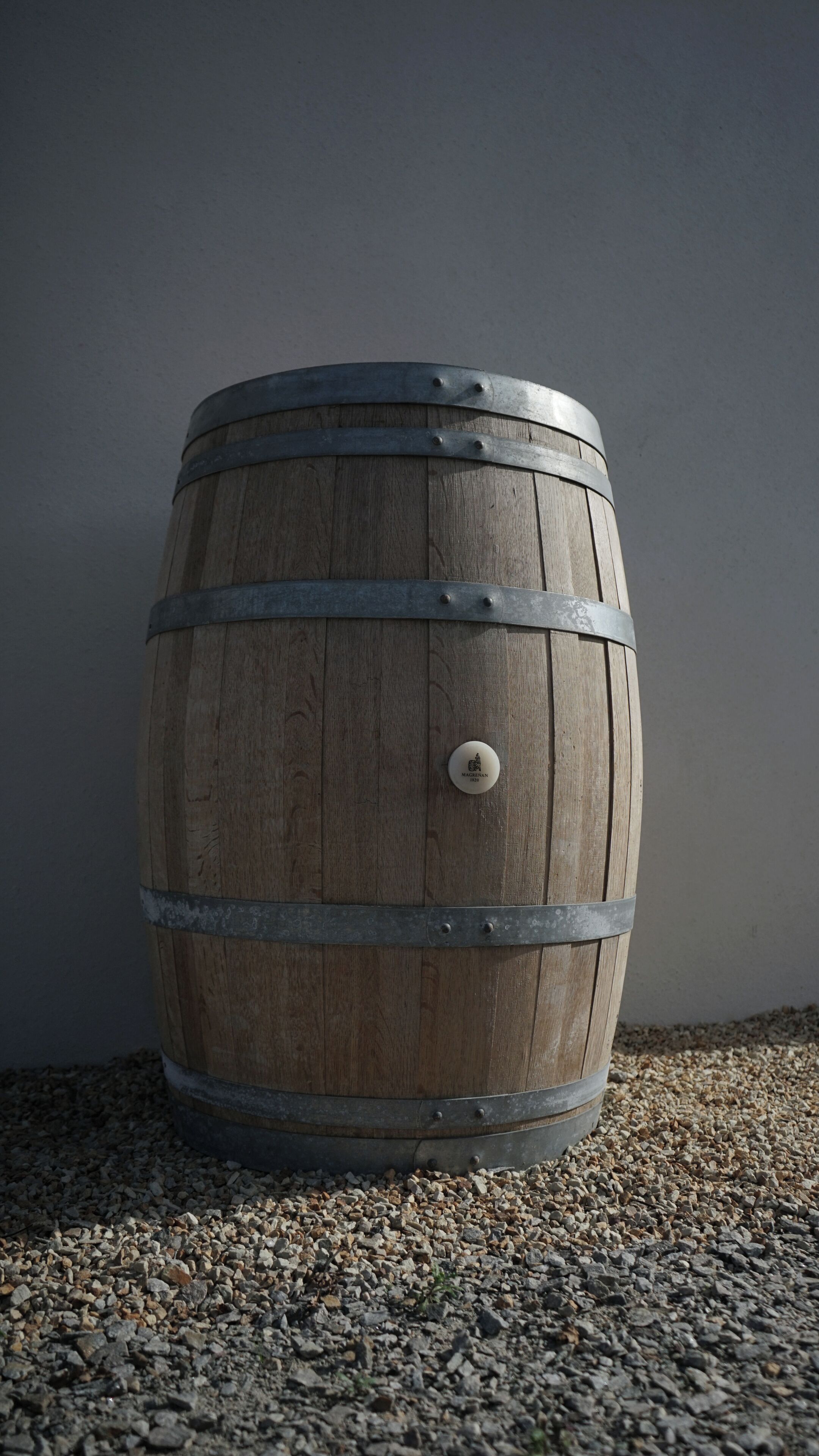 A wooden barrel with metal bands and a knob, placed on a gravel surface against a plain wall.