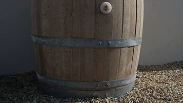 A wooden barrel with metal bands and a knob, placed on a gravel surface against a plain wall.