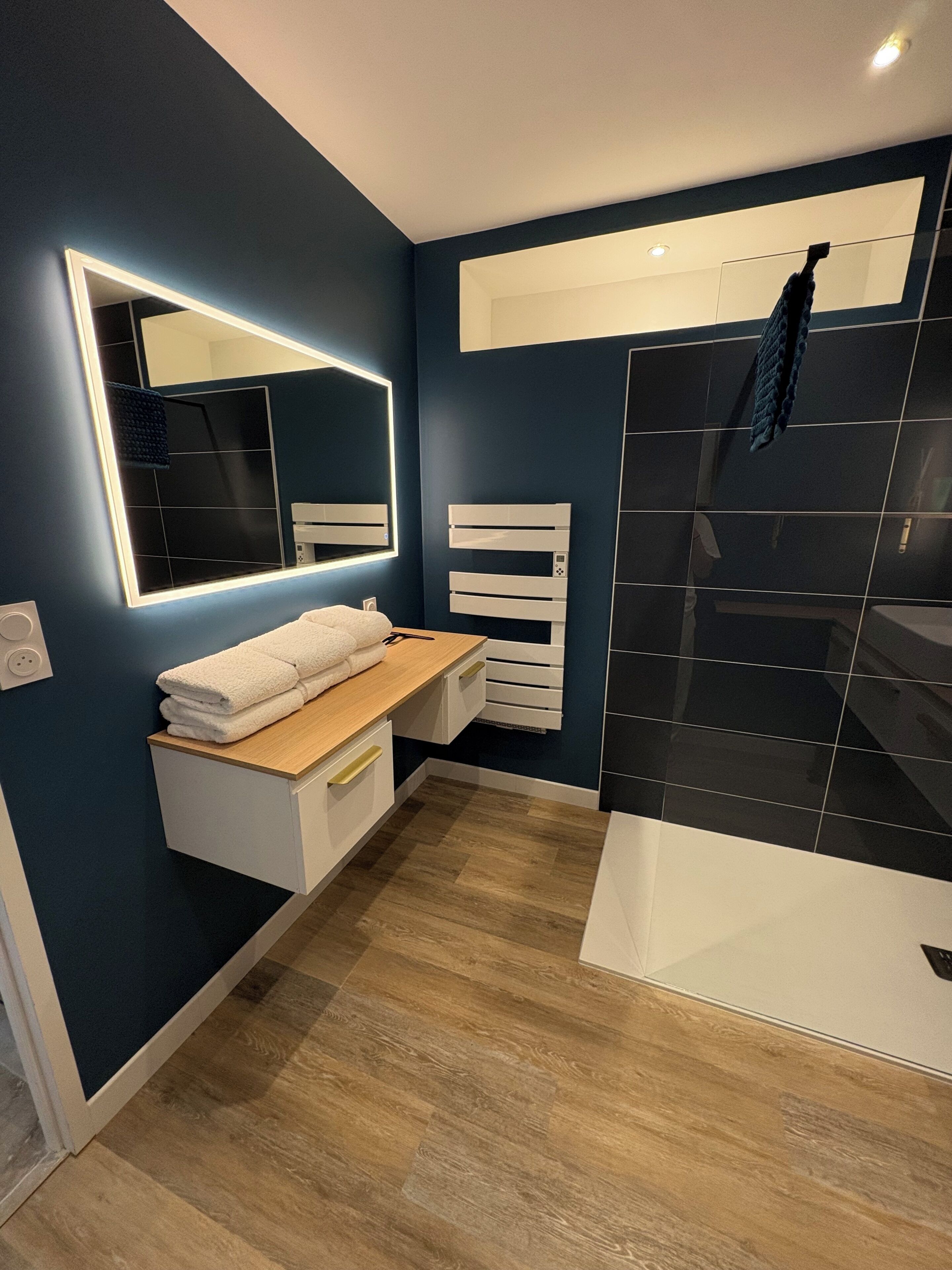 A modern bathroom with a wooden vanity, a large mirror with integrated lighting, a white towel rack, and a black tiled shower enclosure.