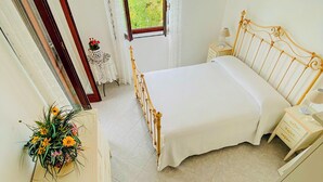 A bedroom with a bed, bedside tables, a dresser, a mirror, and a window with a view of trees. - Villa Rosamaria (Maratea)