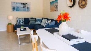 Ein modernes Wohnzimmer mit einem dunkelblauen Sofa, einem weißen Couchtisch und einer Vase mit orangefarbenen Blumen. An der Wand hängen zwei gerahmte Bilder und auf dem Beistelltisch steht eine kleine Pflanze.