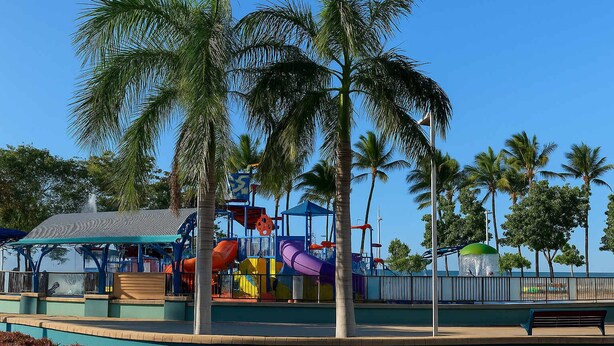 Ein Spielplatz mit Rutschen und Klettergerüsten, umgeben von Palmen und blauem Himmel.
