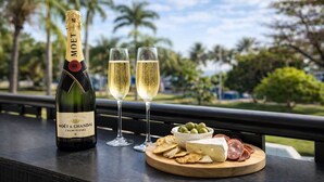 Eine Flasche Moët & Chandon Champagner, zwei Sektgläser und eine Platte mit Käse und Wurst auf einem Balkon mit Blick auf Palmen.