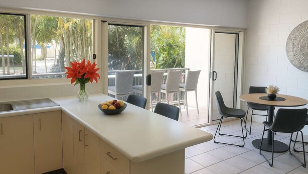 Une cuisine moderne dotée d’un grand îlot central, d’un coin repas avec une table ronde et des chaises, et d’une porte-fenêtre coulissante donnant sur un balcon aménagé avec du mobilier d’extérieur.