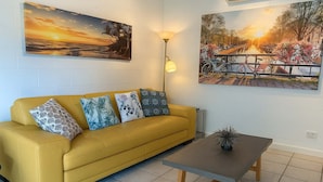 Un canapé jaune agrémenté de coussins à motifs, une petite table sur laquelle est posé un vase, et deux tableaux représentant des paysages, accrochés au mur.