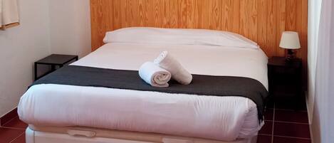 A hotel room with a wooden headboard, a bed with white bedding, a rolled towel, a small nightstand with a lamp, and a suitcase on the floor.