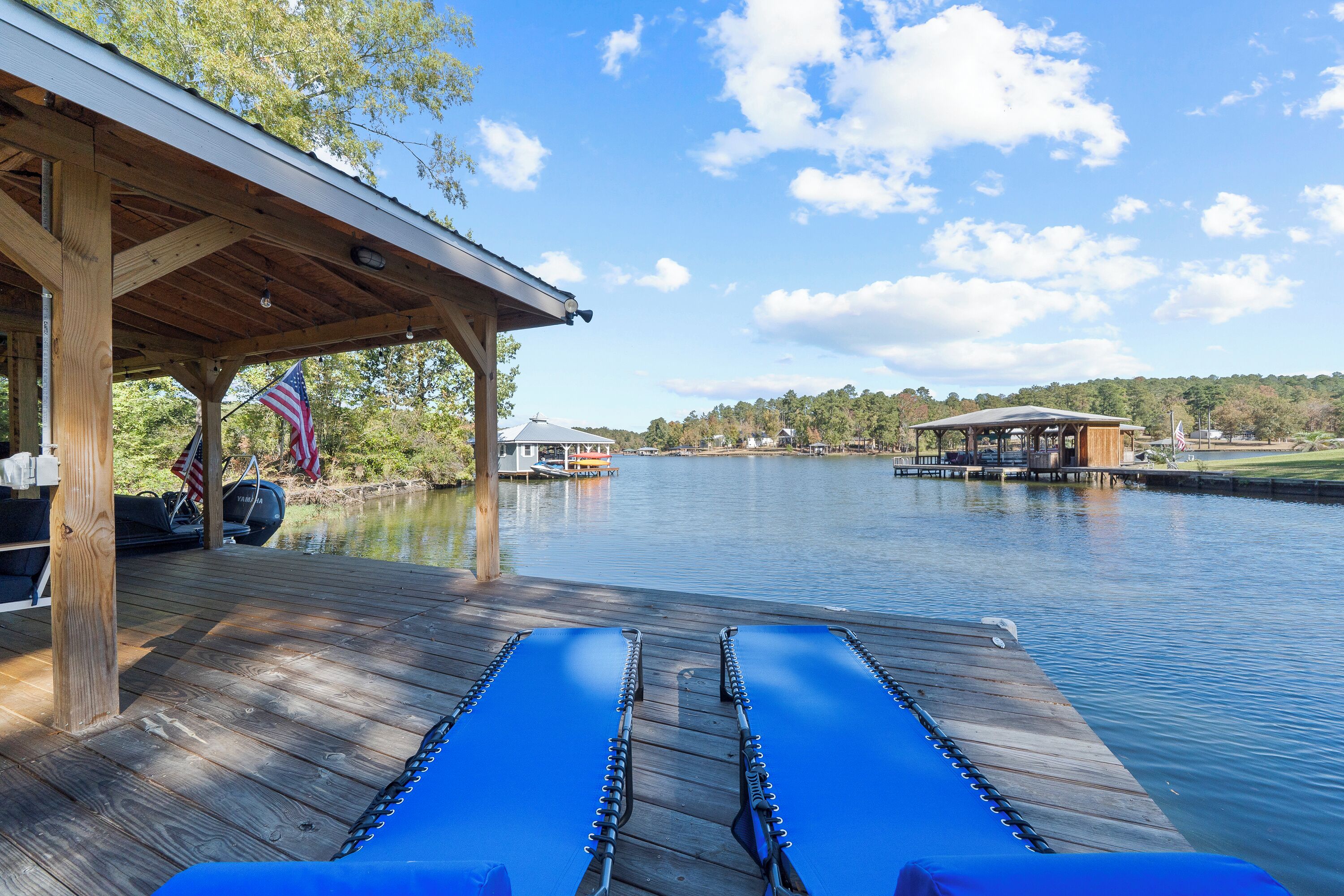 A wooden deck with lounge chairs overlooking a calm lake, a covered area with an American flag, and a distant view of a dock and buildings.