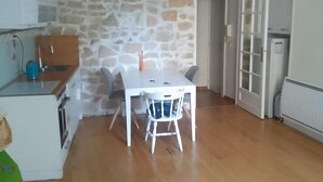 A room with a stone wall, wooden flooring, a white table with chairs, a kitchen area with cabinets and a stove, and a door leading to another room. - Very charming 1 bed room in heart of paris with balcony (Paris)