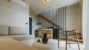 A modern living room with a sofa, ottoman, and a wooden chair. A staircase leads to a kitchen area with wooden cabinets and a black refrigerator. - Orizontas Suites 2 Syvota (Igoumenitsa)