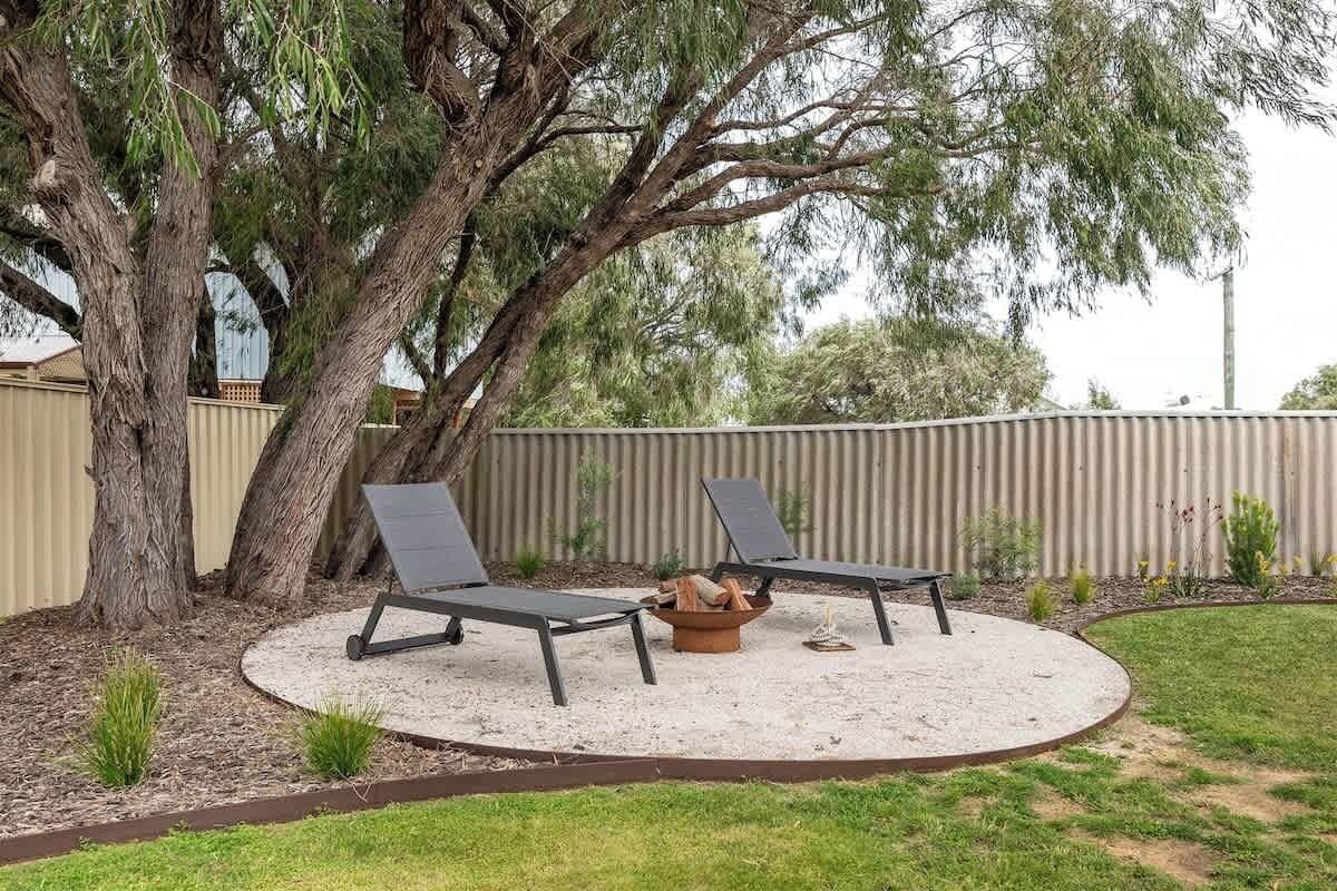 A circular outdoor seating area with two lounge chairs and a fire pit, surrounded by a sandy area and a grassy lawn.