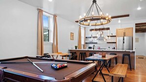 A pool table with a cue stick and balls, a wooden bench, a dining table with chairs, a kitchen area with cabinets and a refrigerator, and a chandelier. - Summit Haus  Cozy Getaway in the Poconos (Pocono Summit)