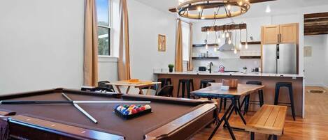 Una mesa de billar con taco y bolas, un banco de madera, una mesa de comedor con sillas, una cocina con alacenas y un refrigerador, y una araña.