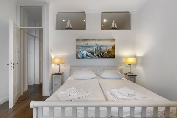 Quarto com cama de casal, duas luminárias de cabeceira, um quadro sobre a cama e uma porta que dá para outro cômodo.