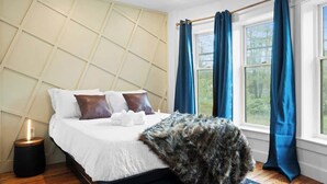Ein Schlafzimmer mit einem Bett, einem Kopfteil, einem Fenster mit Vorhängen und einem Nachttisch mit Lampe.