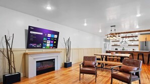 Un salón moderno con televisor de pantalla plana, suelo de madera, sillones de cuero, chimenea y comedor con cocina al fondo.