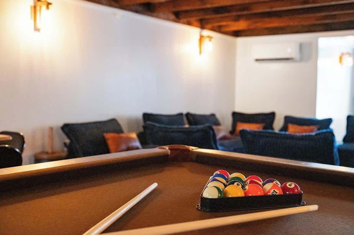 Una mesa de billar con bolas y tacos, un sofá con cojines e iluminación en la pared en una habitación con vigas de madera.