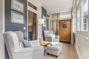 Een modern interieur met twee grijze fauteuils, een klein houten tafeltje en een groot raam met gordijnen.