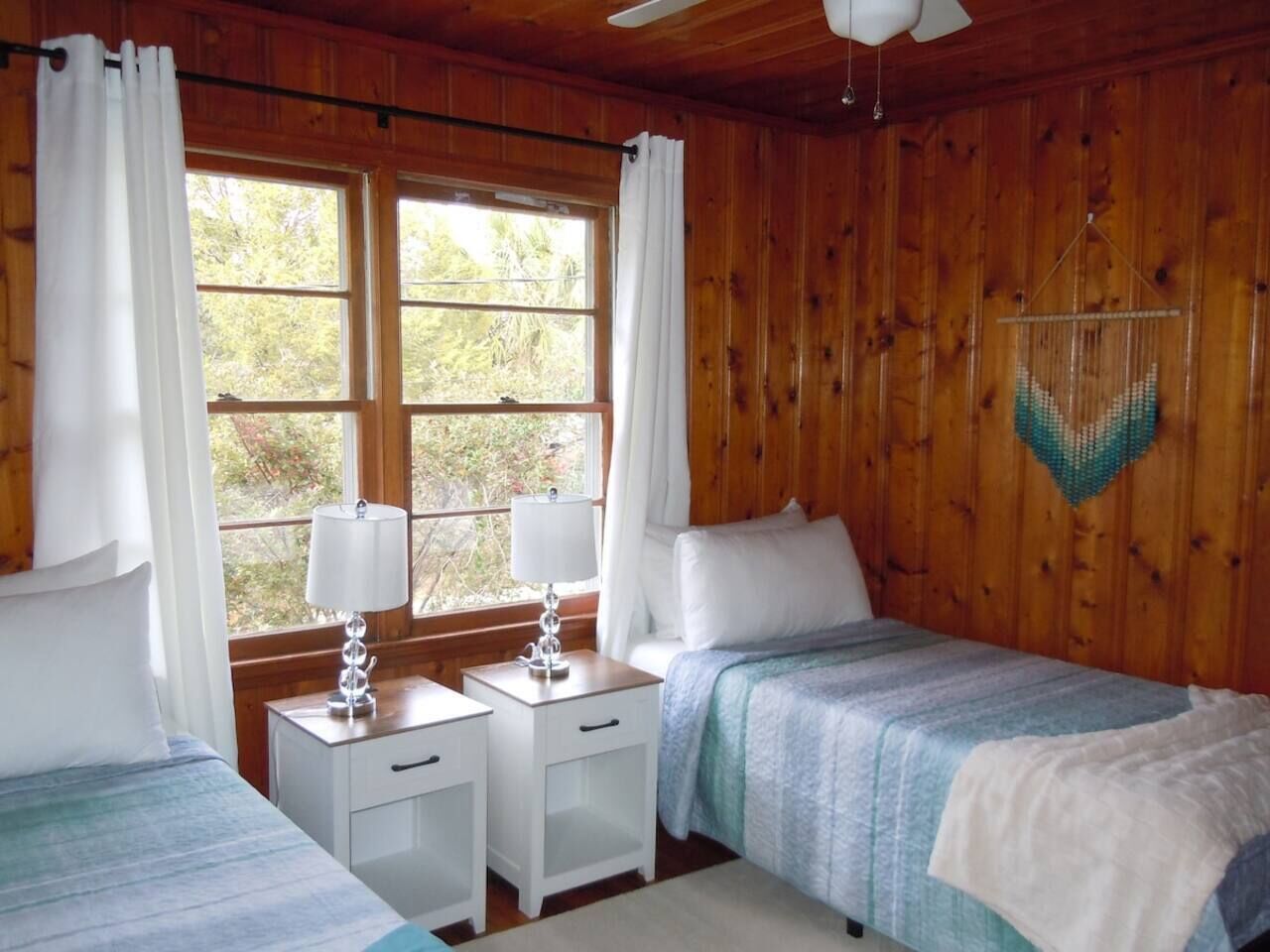 Une chambre de style cabane avec deux lits, des murs en bois, un ventilateur de plafond et une fenêtre avec des rideaux.