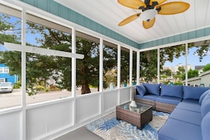 A covered porch with a ceiling fan, large windows, a blue sofa, and a glass coffee table. - 2 Bdrm Beach Cottage- Fire Pit, 3 Blocks to Beach (North Myrtle Beach)