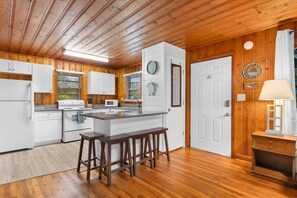 A modern kitchen with wooden flooring, white cabinetry, a central island with stools, and a large window with blinds. - 2 Bdrm Beach Cottage- Fire Pit, 3 Blocks to Beach (North Myrtle Beach)