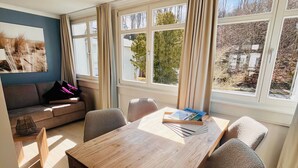 A dining area with a wooden table, four chairs, a sofa, and a large window with curtains. - Apartment "Halbinsel Mönchgut" with Balcony and Wi-Fi (Binz)