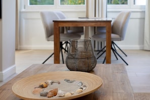 Een houten tafel met een ronde schotel met kiezelstenen en schelpen, een geweven mand, en een eetgedeelte met stoelen en een tafel op de achtergrond.