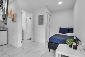 A modern bedroom with a bed, bedside table, and a small kitchenette area. - Tropical Haven | Cozy 1 BR Caribbean Getaway (Miami)