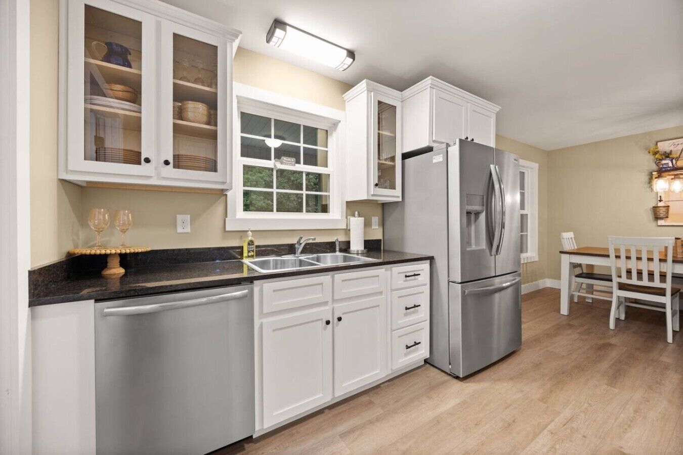 Una cocina con armarios blancos, un refrigerador de acero inoxidable, un lavamanos y una zona de comedor con una mesa y sillas.