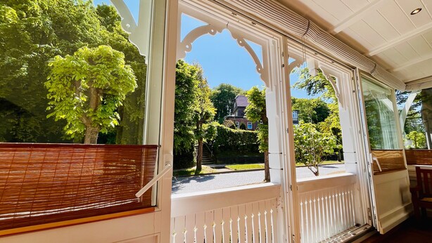 Une véranda couverte, dotée de balustrades blanches et d’un banc en bois, qui donne sur un jardin arboré, avec une maison visible au loin.