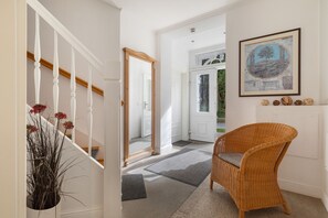 Un pasillo con una escalera blanca, un espejo grande, una silla de mimbre y una obra de arte enmarcada en la pared.
