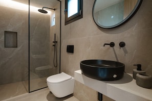 A modern bathroom with a glass-enclosed shower, a white toilet, a black sink, and a round mirror. - Orizontas Suites 1 Syvota (Igoumenitsa)