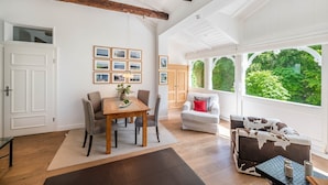 Quarto espaçoso com uma mesa de jantar de madeira, cadeiras, sofá e uma grande janela com vista para o verde.